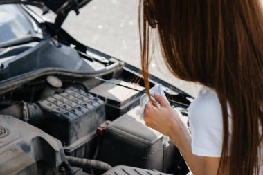 Genç bir kız otoyolun ortasında bozuk bir arabanın yanında duruyor ve motorun yağ seviyesini kontrol ediyor. Aracın bozulması ve arızası. Yardım bekliyorum..