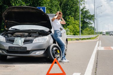 Genç bir kız, otoyolun ortasında kırık bir tekerlekle kırık bir arabanın yanında duruyor ve sıcak bir günde yardım beklerken hayal kırıklığına uğruyor. Arabanın arızası ve arızası. Yardım bekliyorum..