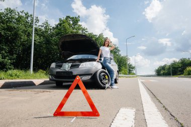 Genç bir kız, otoyolun ortasında kırık bir tekerlekle kırık bir arabanın yanında duruyor ve sıcak bir günde yardım beklerken hayal kırıklığına uğruyor. Arabanın arızası ve arızası. Yardım bekliyorum..
