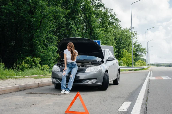 一个年轻的女孩站在高速公路中央一辆抛锚的汽车旁边 打电话求助 汽车故障和抛锚 等人帮忙 — 图库照片