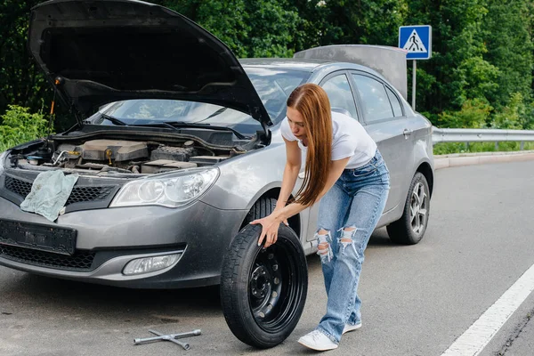 A young girl stands near a broken car in the middle of the highway and tries to change a broken wheel on a hot sunny day. Failure and breakdown of the car. Waiting for help.
