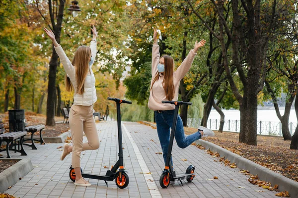 マスクの2人の若い美しい女の子は 暖かい秋の日に公園の電動スクーターに乗っています 公園を歩く — ストック写真