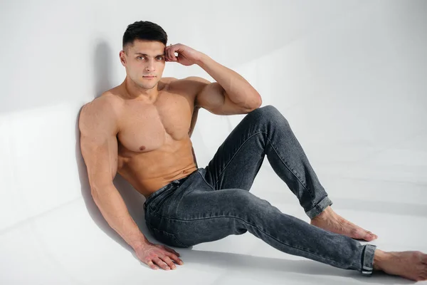 A young sexy athlete with perfect abs sits on the floor in the studio topless in jeans in the background. Healthy lifestyle, proper nutrition, training programs and nutrition for weight loss.