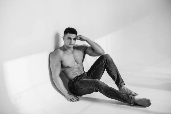A young sexy athlete with perfect abs is sitting in the studio topless in jeans. Healthy lifestyle, proper nutrition, training programs and nutrition for weight loss. Black and white.