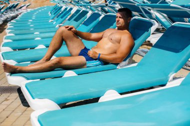 Genç bir atletik adam güneşli bir günde güneşlenirken mutlu bir şekilde gülümsüyor ve güneşleniyor. İyi tatiller. Yaz tatilleri ve turizm.