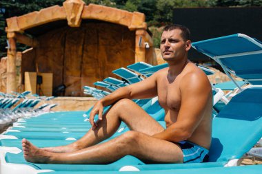 Genç bir atletik adam güneşli bir günde güneşlenirken mutlu bir şekilde gülümsüyor ve güneşleniyor. İyi tatiller. Yaz tatilleri ve turizm.