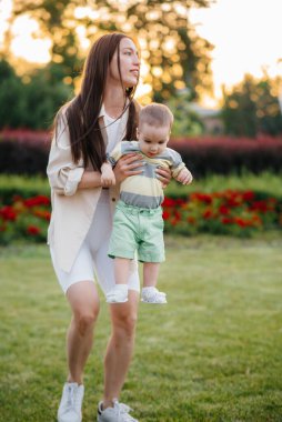 Genç ve tatlı bir anne, küçük oğluna gün batımında çimlerin üzerinde ilk adımlarını atması için yardım eder ve öğretir..