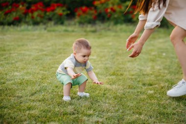 Genç ve tatlı bir anne, küçük oğluna gün batımında çimlerin üzerinde ilk adımlarını atması için yardım eder ve öğretir..