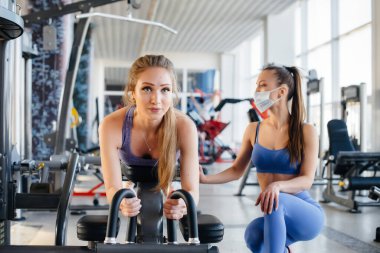 Salgın sırasında maske takan iki güzel kız spor salonunda egzersiz yapıyor. Halka açık yerlerde sosyal mesafeler.