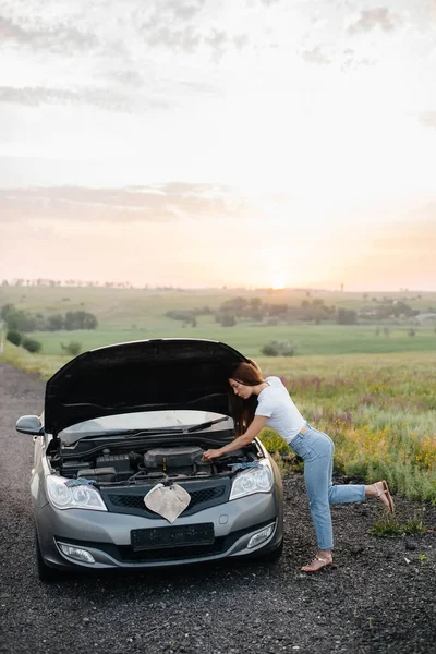 日落时分 一个小女孩站在公路中央一辆破车的旁边 试图修理它 汽车的故障和修理 问题的解决 — 图库照片