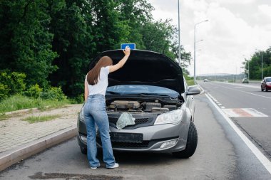Genç bir kız otoyolun ortasında bozuk bir arabanın yanında duruyor ve kaputun altına bakıyor. Aracın bozulması ve arızası. Yardım bekliyorum..