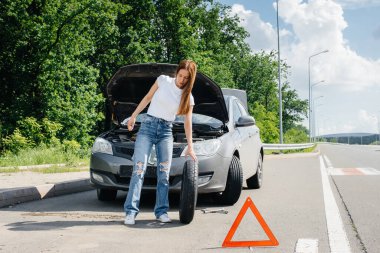 Genç bir kız otoyolun ortasında bozuk bir arabanın yanında duruyor ve güneşli bir günde kırık bir tekerleği değiştirmeye çalışıyor. Aracın bozulması ve arızası. Yardım bekliyorum..