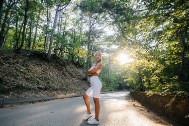 Genç ve güzel bir kız antrenmandan önce poz veriyor, sık bir ormanda, gün batımında. Sağlıklı bir yaşam tarzı ve temiz havada koşmak..