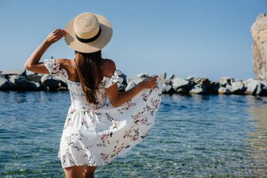 Şapkalı ve sırtında açık renk elbisesi olan güzel bir kadın güneşli bir günde okyanus kıyısında büyük kayaların arkasında yürüyor. Turizm ve tatil gezisi.