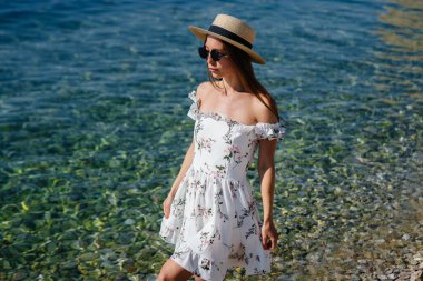 Şapkalı, gözlüklü ve açık renk elbiseli güzel bir kadın güneşli bir günde okyanus kıyısında büyük kayaların arkasında yürüyor. Turizm ve turizm gezileri.