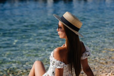 Şapkalı, gözlüklü ve açık renk elbiseli güzel bir kadın güneşli bir günde okyanus kıyısında büyük kayaların arkasında oturuyor. Turizm ve turizm gezileri.