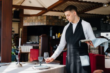 Şık üniformalı genç bir erkek garson güzel bir gurme restoranında masaya servis yapmakla meşgul. Restoran faaliyetleri, en üst düzeyde..