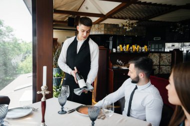 Şık önlüklü genç bir garson bir restoranda güzel bir çifte güzel bir şarap ikram ediyor. Müşteri hizmetleri.