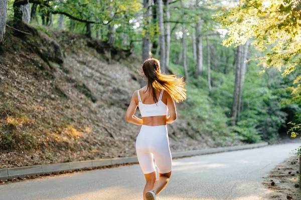 日落时分 一个身穿白色运动服的年轻漂亮的姑娘背着她在茂密的森林里的路上奔跑着 在新鲜空气中做运动 健康的生活方式 — 图库照片