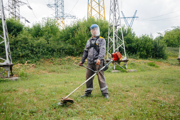 Молодой человек косит траву на территории электрической подстанции в комбинезоне. Очистка травы на предприятии, реализация мероприятий по пожарной безопасности