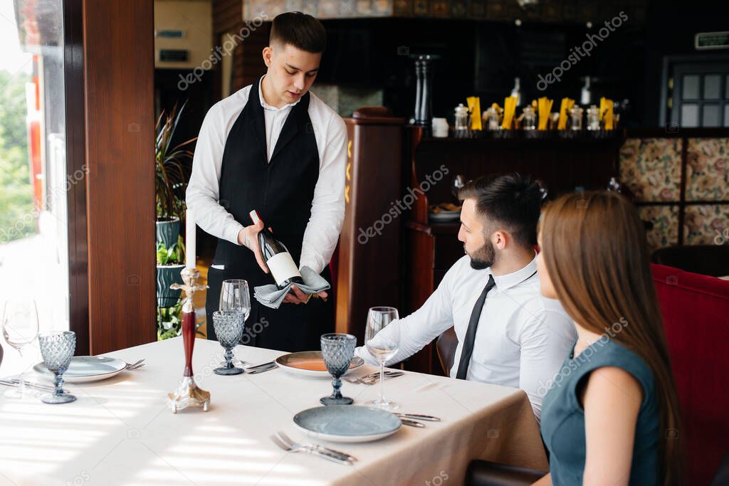 A young waiter in a stylish apron demonstrates and offers a fine wine to a beautiful couple in a restaurant. Customer service.