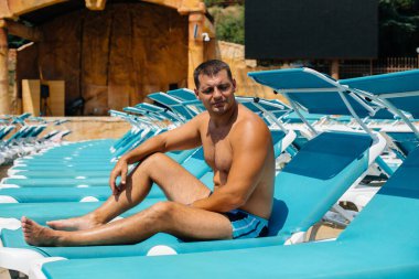 Genç bir atletik adam güneşli bir günde güneşlenirken mutlu bir şekilde gülümsüyor ve güneşleniyor. İyi tatiller. Yaz tatilleri ve turizm.
