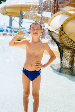 Yedi yaşında mutlu bir çocuk gülümser ve su parkındaki slaytların arka planına karşı bir sınıf gösterir. İyi tatiller. Yaz tatilleri ve turizm.