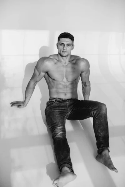 A young sexy athlete with perfect abs is sitting in the studio topless in jeans. Healthy lifestyle, proper nutrition, training programs and nutrition for weight loss. Black and white.