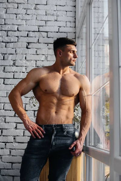 A young sexy athlete with perfect abs poses near the window in the studio topless in jeans on the background. Healthy lifestyle, proper nutrition, training programs and nutrition for weight loss.