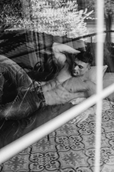 A young sexy athlete with perfect abs is lying on the bed in the studio topless in jeans. Healthy lifestyle, proper nutrition, training programs and nutrition for weight loss. Black and white.
