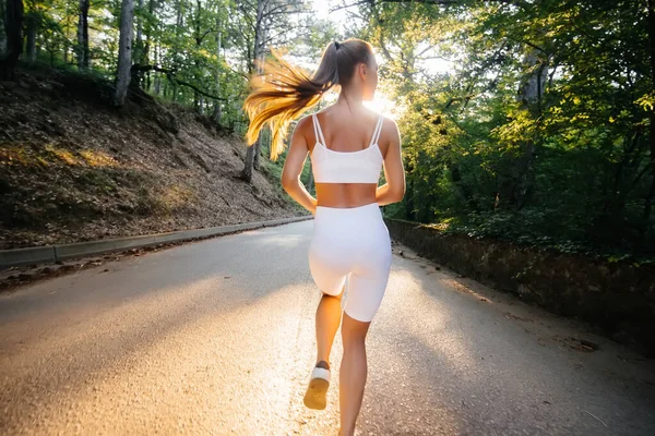 白いスポーツ服を着た若い美しい少女が 日没の間 鬱蒼とした森の中の道路で彼女と一緒に走っています 新鮮な空気の中でスポーツをしている 健康的なライフスタイル — ストック写真