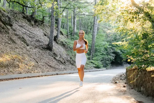 落日时分 一个身穿白色运动服的年轻漂亮的姑娘在茂密的森林里的路上跑着 在新鲜空气中做运动 健康的生活方式 — 图库照片