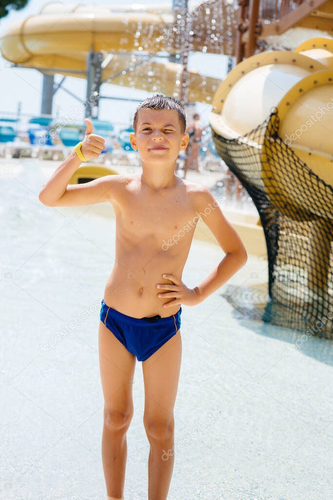 A happy boy of seven years old smiles and shows a class against the background of slides in a water park. Happy vacation vacation. Summer holidays and tourism.