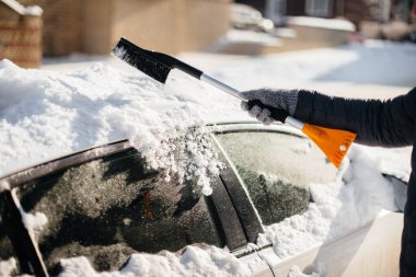 Genç bir adam güneşli, soğuk bir günde kar yağdıktan sonra arabasını temizler..