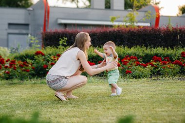 Genç ve tatlı bir anne, küçük oğluna gün batımında çimlerin üzerinde ilk adımlarını atması için yardım eder ve öğretir..