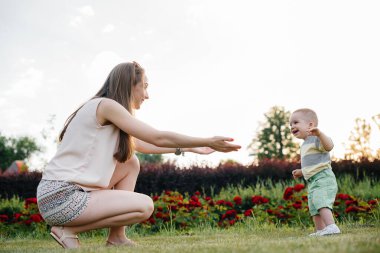 Genç ve tatlı bir anne, küçük oğluna gün batımında çimlerin üzerinde ilk adımlarını atması için yardım eder ve öğretir..