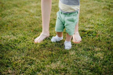 Genç ve tatlı bir anne, küçük oğluna gün batımında çimlerin üzerinde ilk adımlarını atması için yardım eder ve öğretir..