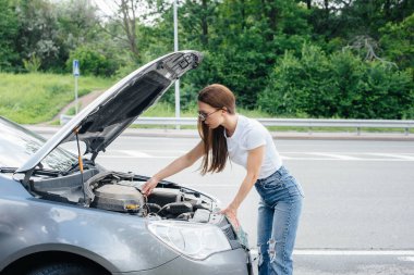 Genç bir kız otoyolun ortasında bozuk bir arabanın yanında duruyor ve kaputun altına bakıyor. Aracın bozulması ve arızası. Yardım bekliyorum..