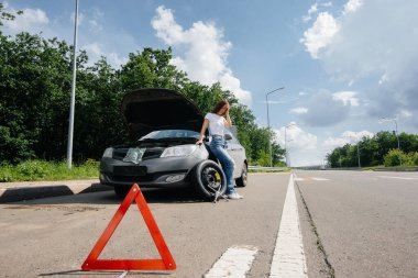 A young girl stands near a broken car with a broken wheel in the middle of the highway and is frustrated waiting for help on a hot day. Breakdown and breakdown of the car. Waiting for help. clipart