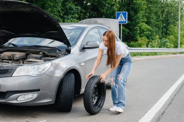 Genç bir kız otoyolun ortasında bozuk bir arabanın yanında duruyor ve güneşli bir günde kırık bir tekerleği değiştirmeye çalışıyor. Aracın bozulması ve arızası. Yardım bekliyorum..