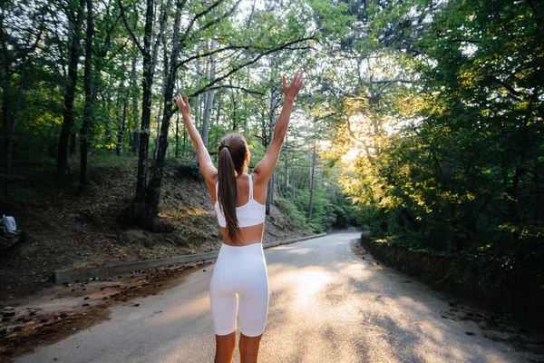 若い美しい少女は 日没の間 鬱蒼とした森の中の道路上で トレーニングを実行する前に彼女の背中を上げてポーズをとります 健康的なライフスタイルと新鮮な空気の中で実行している — ストック写真