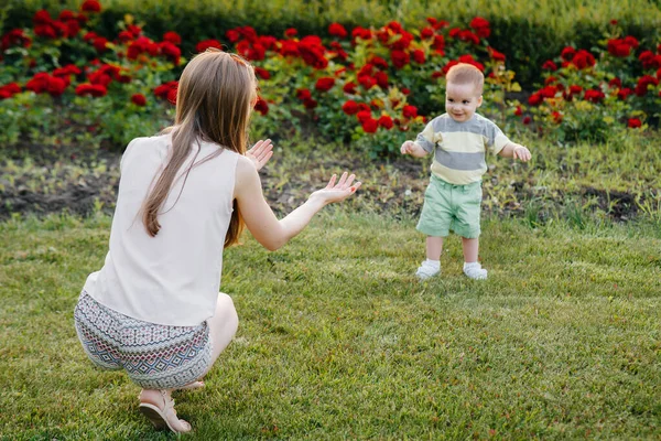Genç ve tatlı bir anne, küçük oğluna gün batımında çimlerin üzerinde ilk adımlarını atması için yardım eder ve öğretir..