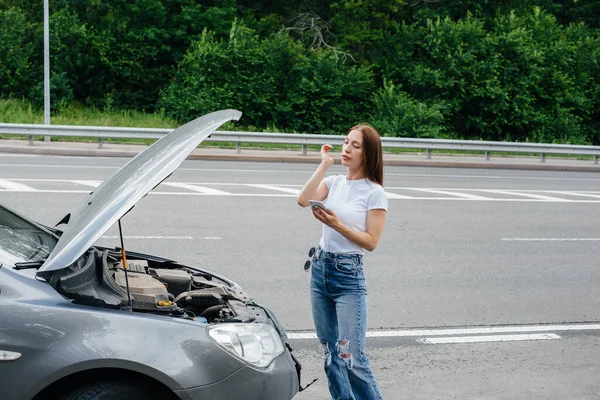 一个年轻的女孩站在高速公路中央一辆抛锚的汽车旁边 打电话求助 汽车故障和抛锚 等人帮忙 — 图库照片