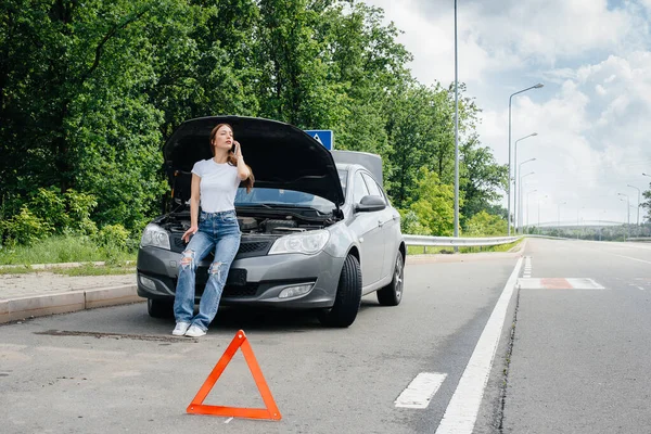 一个年轻的女孩站在高速公路中央一辆抛锚的汽车旁边 打电话求助 汽车故障和抛锚 等人帮忙 — 图库照片