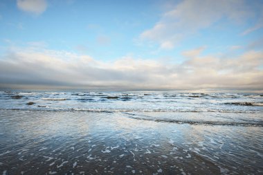 Gün batımında karla kaplı Baltık deniz kıyısından bir manzara. Riga Körfezi, Letonya. Dramatik bir gökyüzü. Kristal berrak mavi su. Kararsız hava, dalgalar ve su sıçramaları. Kış turizmi, küresel ısınma teması