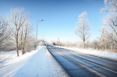 Temizlikten sonra boş bir asfalt yol. Sokak fenerleri yakın plan. Taze karda araba izleri. Arka planda karla kaplı huş ağacı ormanı var. Açık mavi gökyüzü. Finlandiya 'da kış gezisi. Genel ısınma teması