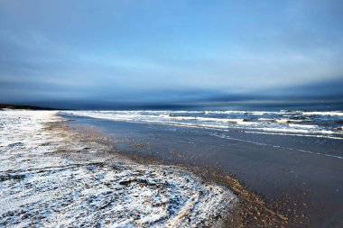 Gün batımında karla kaplı Baltık deniz kıyısından bir manzara. Riga Körfezi, Letonya. Dramatik mavi gökyüzü. Kararsız hava, dalgalar ve su sıçramaları. Kış turizmi, küresel ısınma teması