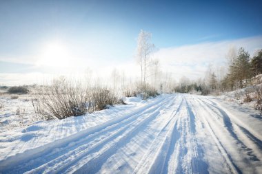 Güneşli bir günde kırsal alanda karla kaplı tek şeritli yol. Finlandiya. Açık mavi gökyüzü. Yaprak döken ve yemyeşil ağaçlar arka planda kırağı kaplamış. Idyllic kış manzarası