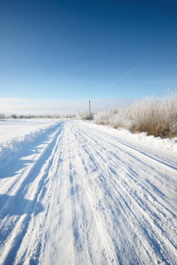 Güneşli bir günde kırsal alanda karla kaplı tek şeritli yol. Finlandiya. Açık mavi gökyüzü. Yaprak döken ve yemyeşil ağaçlar arka planda kırağı kaplamış. Idyllic kış manzarası