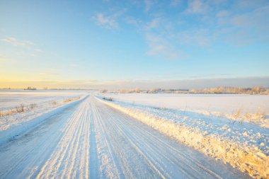 Gün batımında kar fırtınasından sonra karla kaplı arazide köy yolu. Gökyüzü açık, altın ışık. Idyllic kırsal kesim. Panoramik manzara. Noel, lojistik, tehlikeli sürüş, off-road, ulaşım